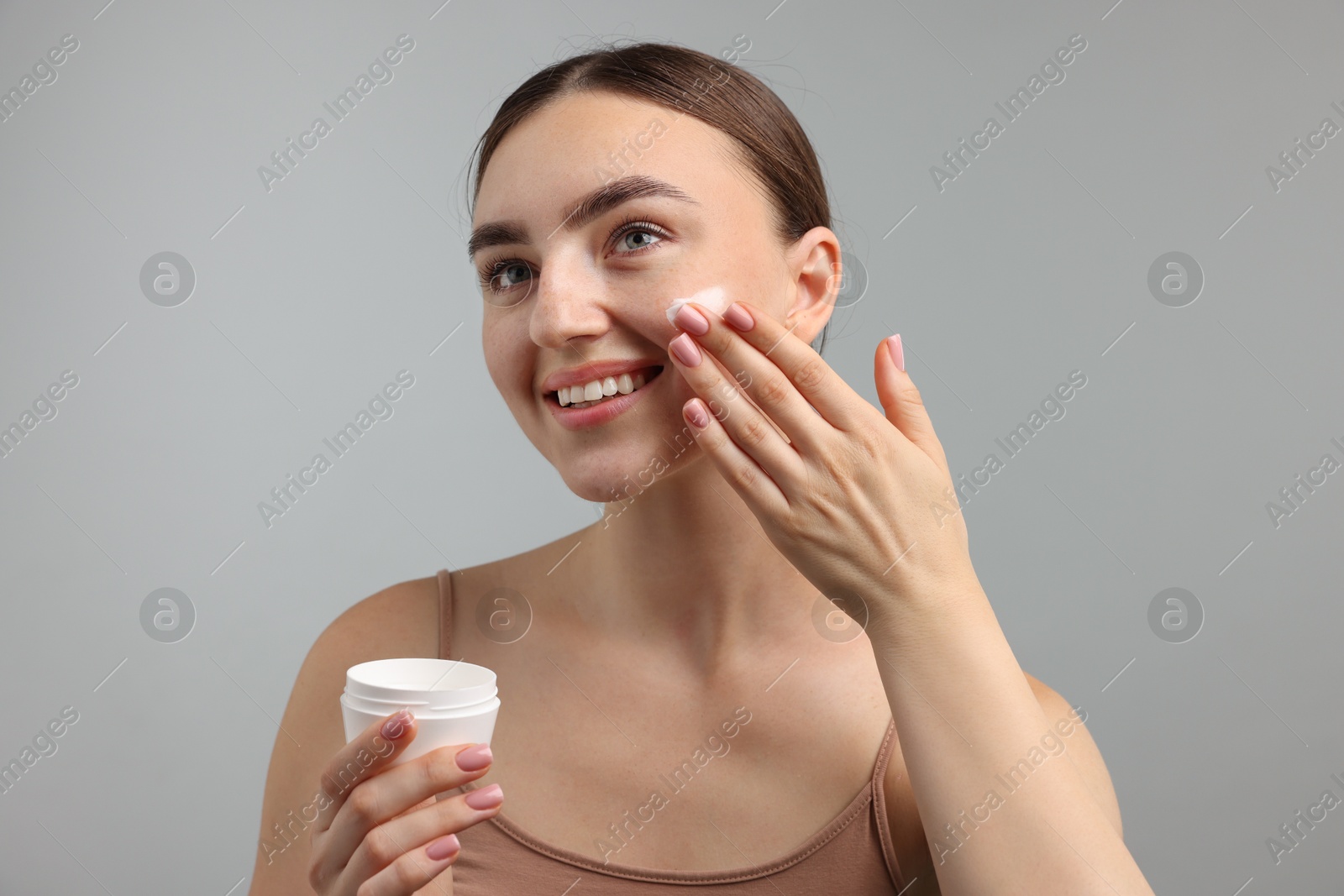 Photo of Beautiful woman applying cream onto her face on gray background