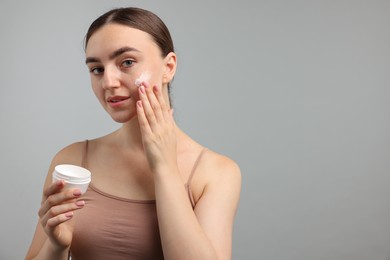 Photo of Beautiful woman applying cream onto her face on gray background