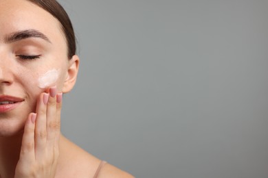 Photo of Beautiful woman applying cream onto her face on gray background, closeup. Space for text