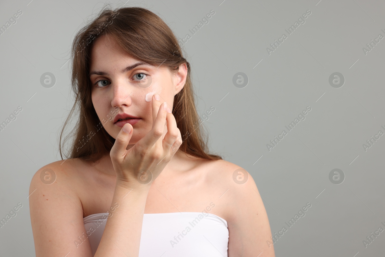 Photo of Beautiful woman applying cream onto her face on gray background. Space for text