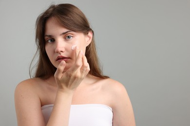 Photo of Beautiful woman applying cream onto her face on gray background. Space for text