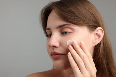 Photo of Beautiful woman applying cream onto her face on gray background, closeup. Space for text