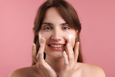 Photo of Beautiful woman applying cream onto her face on pink background, closeup