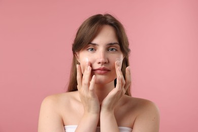 Photo of Beautiful woman applying cream onto her face on pink background