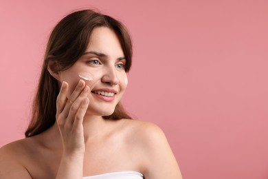 Photo of Beautiful woman applying cream onto her face on pink background. Space for text