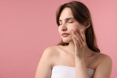 Photo of Beautiful woman applying cream onto her face on pink background. Space for text