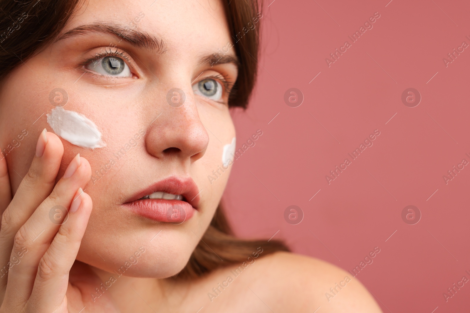 Photo of Beautiful woman applying cream onto her face on pink background, closeup. Space for text