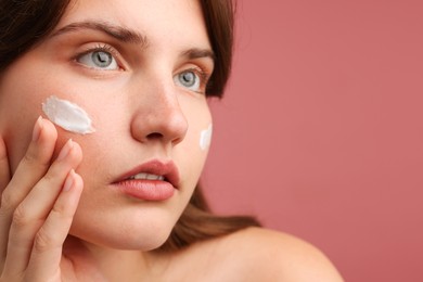 Photo of Beautiful woman applying cream onto her face on pink background, closeup. Space for text