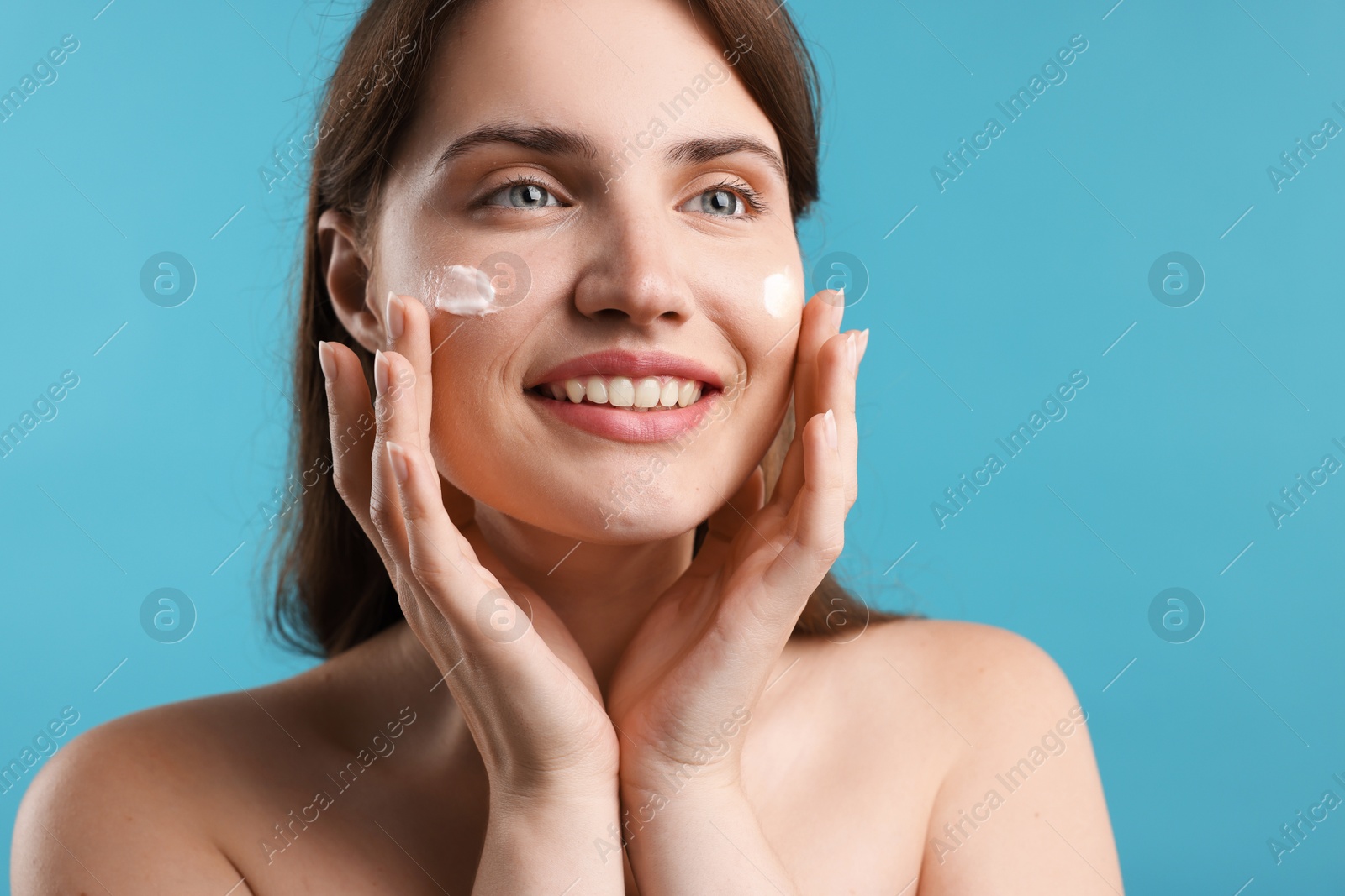 Photo of Beautiful woman applying cream onto her face on light blue background, closeup