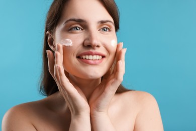 Photo of Beautiful woman applying cream onto her face on light blue background, closeup