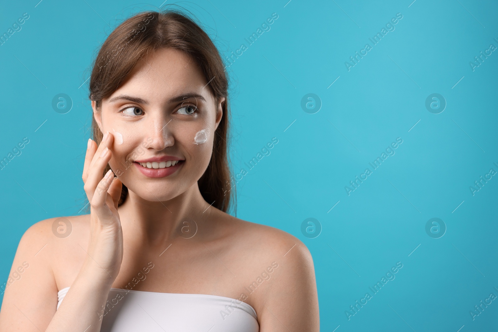 Photo of Beautiful woman applying cream onto her face on light blue background. Space for text
