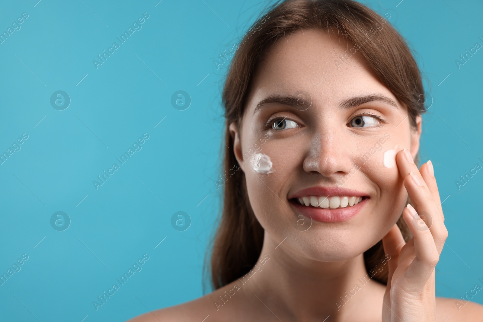 Photo of Beautiful woman applying cream onto her face on light blue background, closeup. Space for text