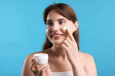 Photo of Beautiful woman applying cream onto her face on light blue background