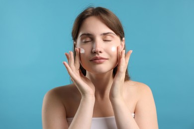 Photo of Beautiful woman applying cream onto her face on light blue background