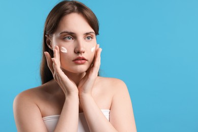 Photo of Beautiful woman applying cream onto her face on light blue background. Space for text
