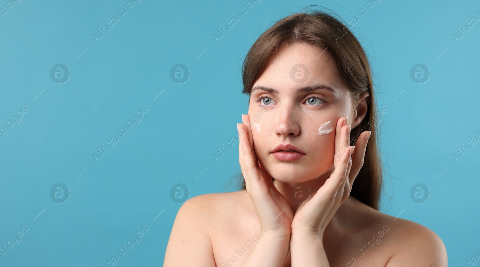 Photo of Beautiful woman applying cream onto her face on light blue background. Space for text