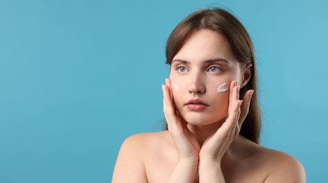 Photo of Beautiful woman applying cream onto her face on light blue background. Space for text