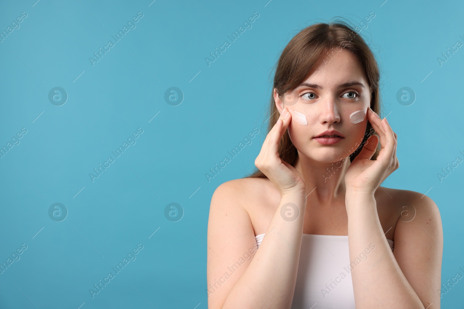 Photo of Beautiful woman applying cream onto her face on light blue background. Space for text