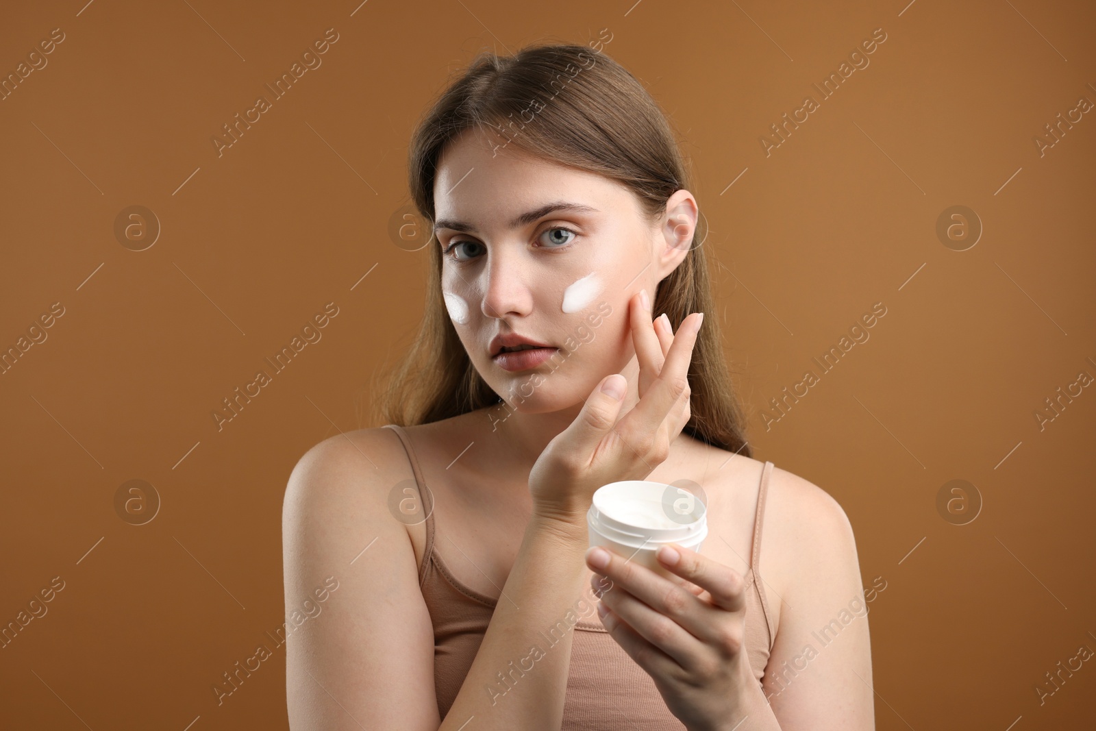 Photo of Beautiful woman applying cream onto her face on brown background