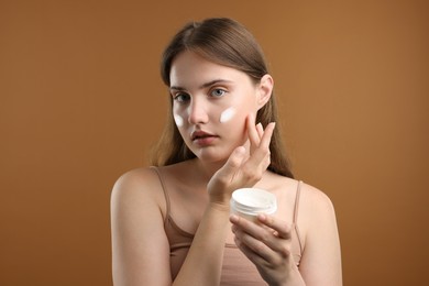 Photo of Beautiful woman applying cream onto her face on brown background