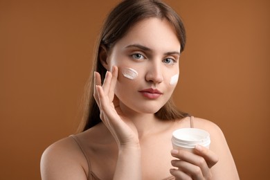 Photo of Beautiful woman applying cream onto her face on brown background