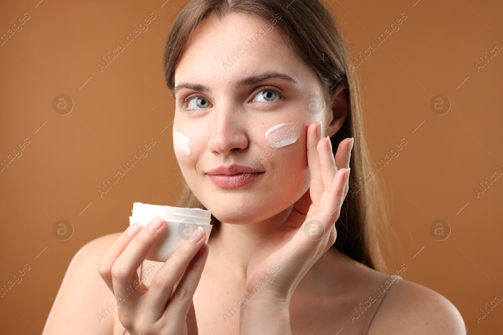 Photo of Beautiful woman applying cream onto her face on brown background