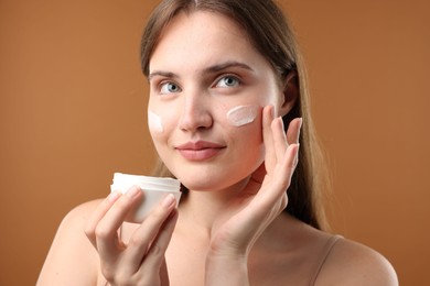 Photo of Beautiful woman applying cream onto her face on brown background