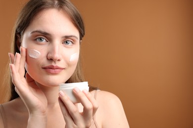 Photo of Beautiful woman applying cream onto her face on brown background. Space for text