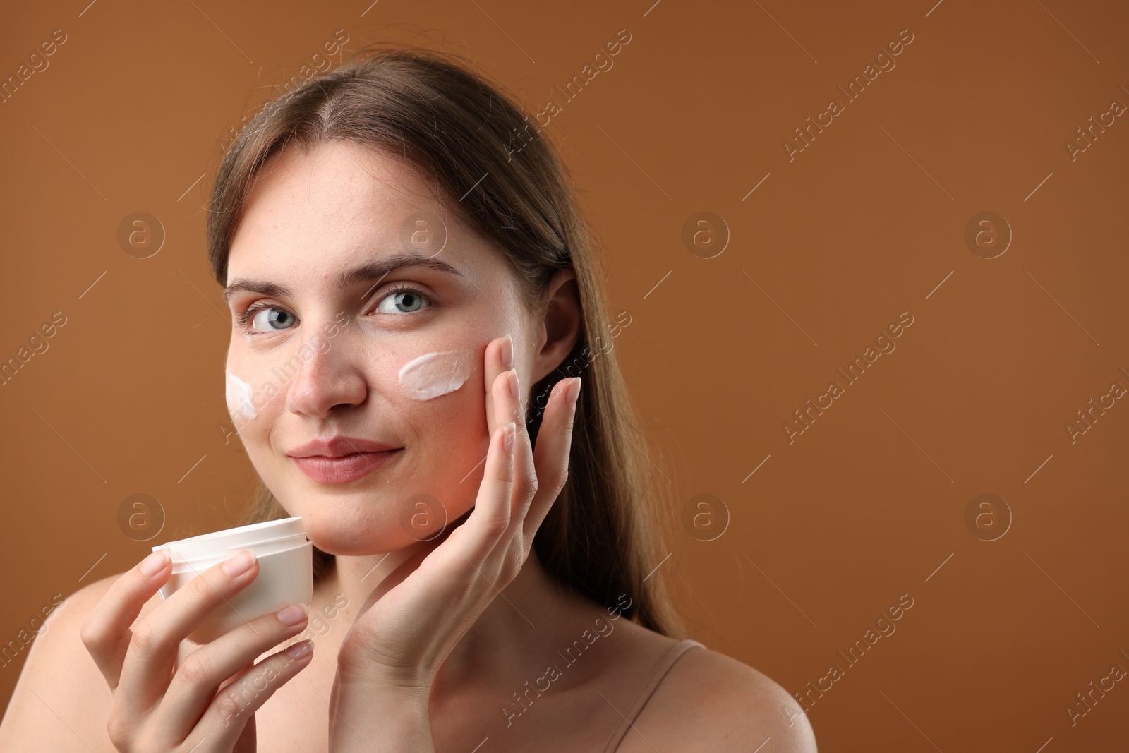 Photo of Beautiful woman applying cream onto her face on brown background. Space for text