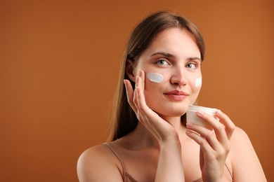 Photo of Beautiful woman applying cream onto her face on brown background. Space for text