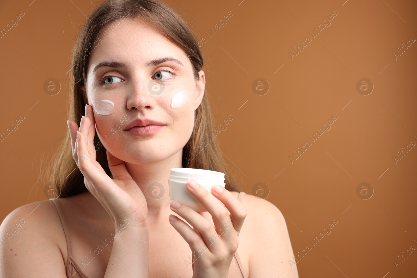 Photo of Beautiful woman applying cream onto her face on brown background. Space for text