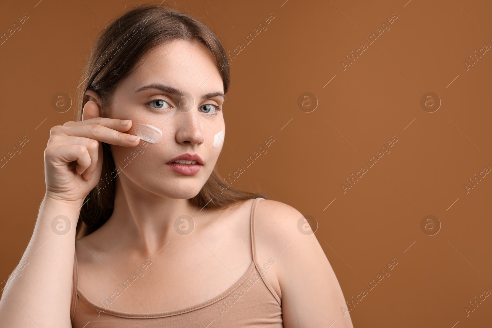 Beautiful woman applying cream onto her face on brown background. Space for text Photo of Beautiful woman applying cream onto her face on brown background. Space for text