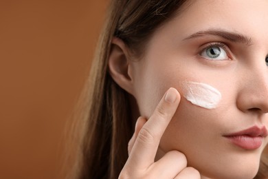 Photo of Woman applying cream onto her face on brown background, closeup