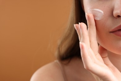 Photo of Woman applying cream onto her face on brown background, closeup. Space for text