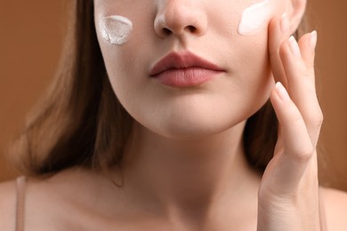 Photo of Woman applying cream onto her face on brown background, closeup