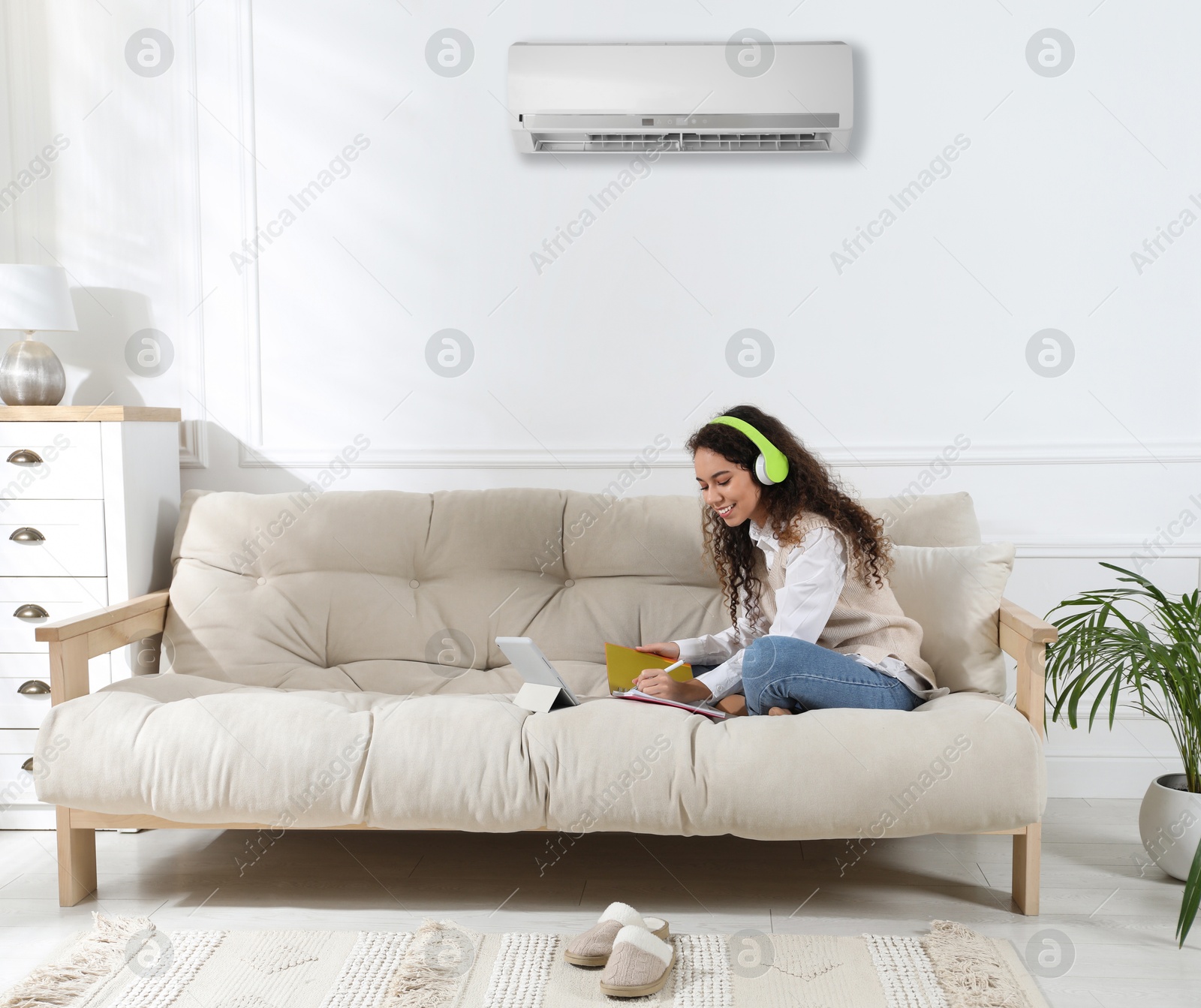 Image of Woman with headphones and tablet studying on sofa at home. Air conditioner keeping comfortable temperature in room