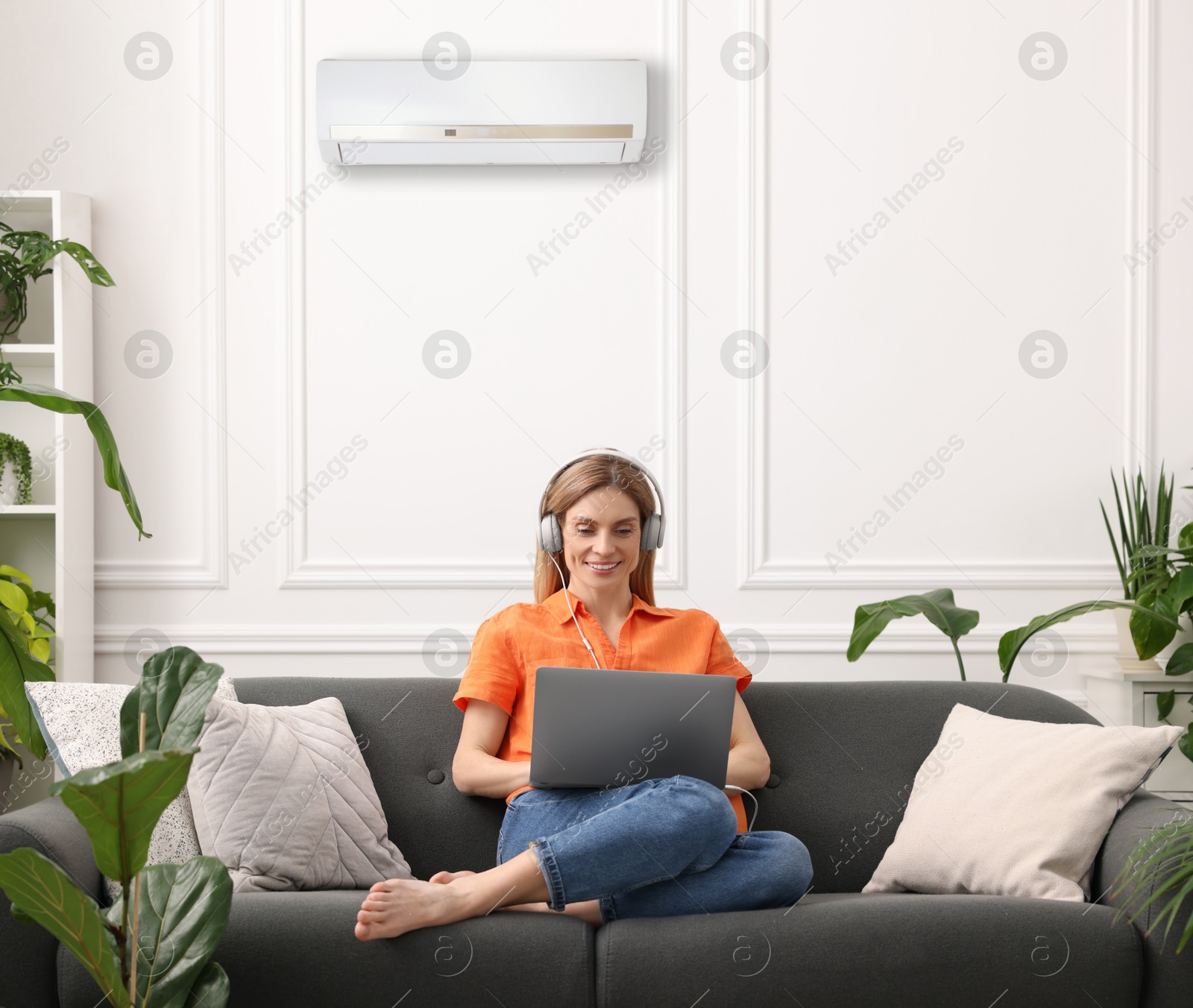Image of Woman in headphones using laptop on sofa at home. Air conditioner keeping comfortable temperature in room