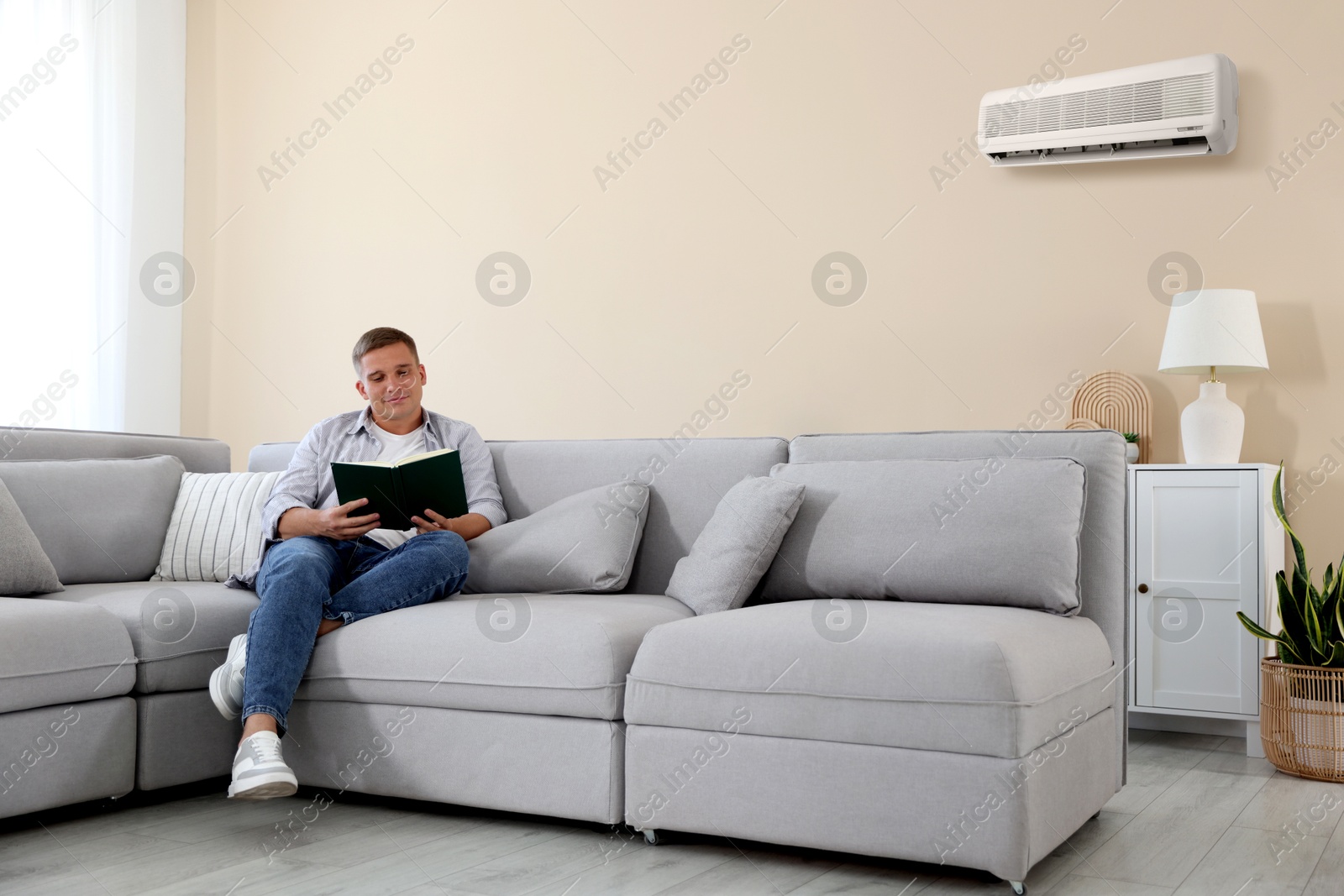 Image of Young man reading book on sofa at home. Air conditioner keeping comfortable temperature in room