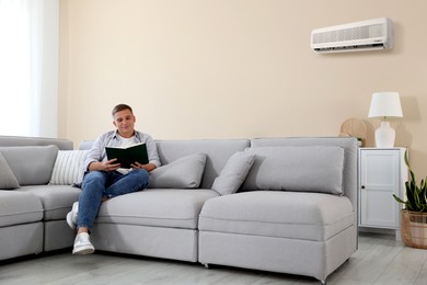 Image of Young man reading book on sofa at home. Air conditioner keeping comfortable temperature in room