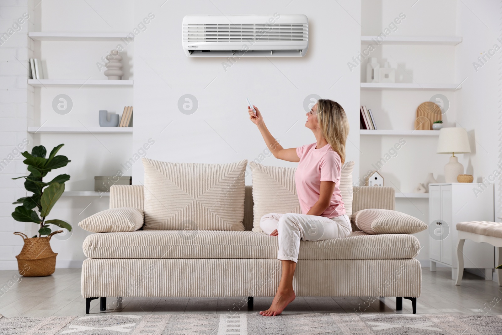 Image of Woman using remote to control air conditioner on sofa at home