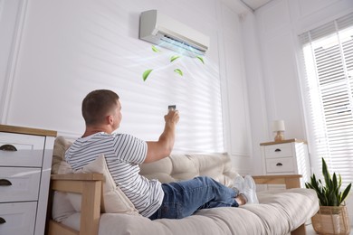 Image of Man using remote to control air conditioner on sofa at home, low angle view