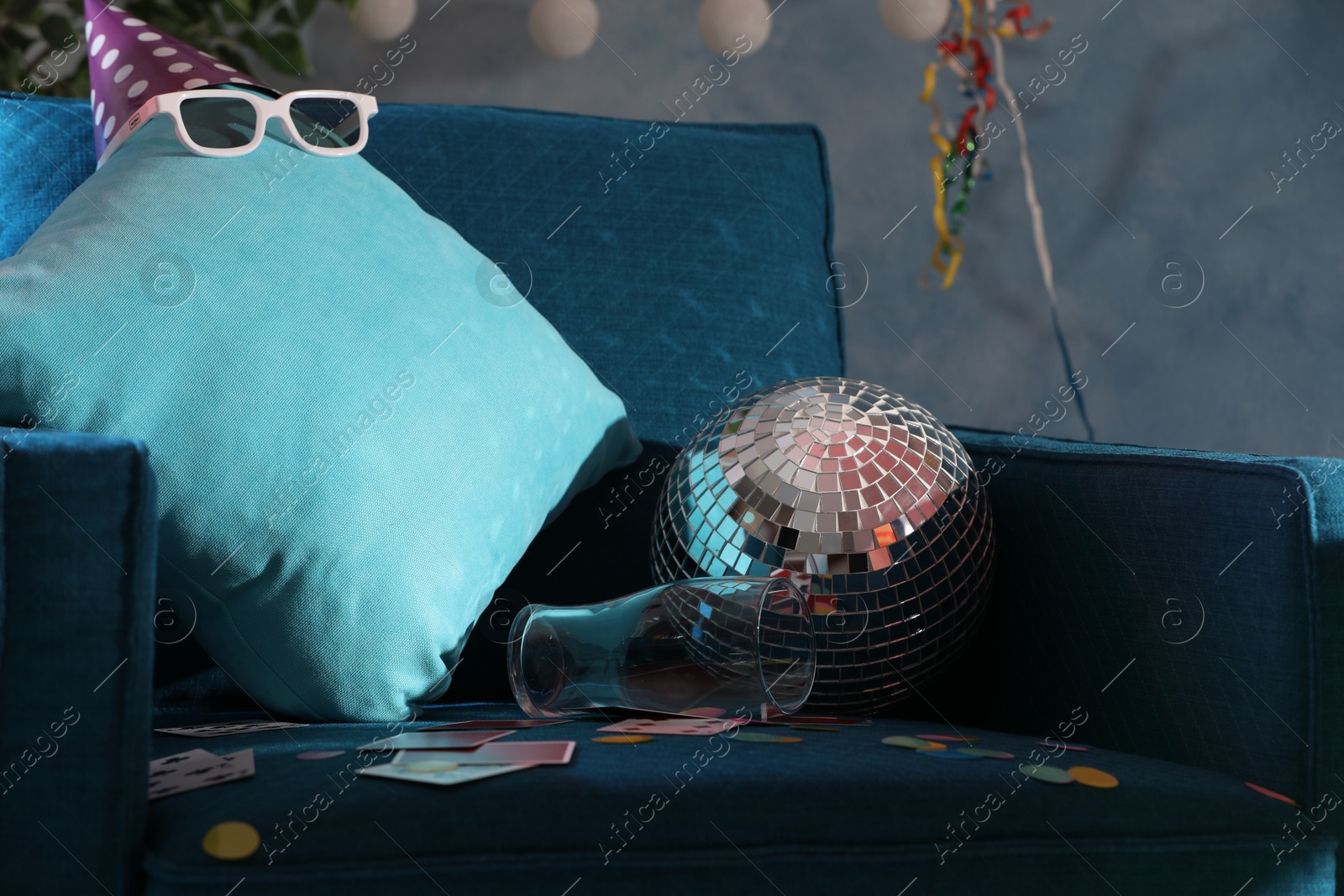 Photo of Disco ball, empty glass and cushion on armchair in messy room after party