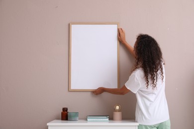 Photo of African American woman hanging empty frame on pale rose wall over table in room, back view. Mockup for design