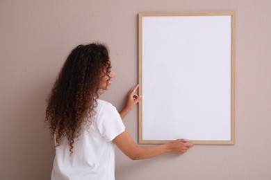 Photo of African American woman hanging empty frame on pale rose wall, back view. Mockup for design