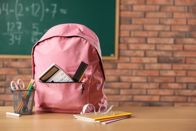 Photo of Pink backpack with different stationery on desk in classroom. Space for text