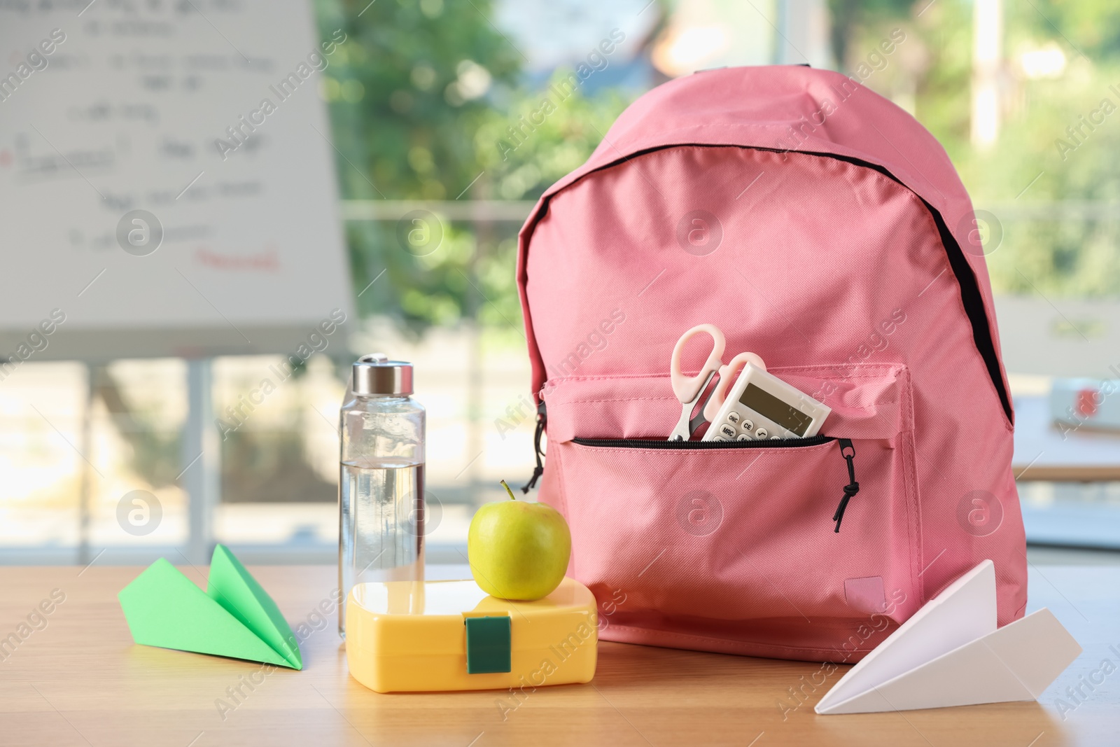Photo of Backpack with different stationery, lunchbox, apple, bottle of water and paper planes on desk in classroom