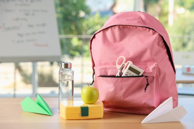 Photo of Backpack with different stationery, lunchbox, apple, bottle of water and paper planes on desk in classroom