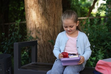 Photo of Cute little girl holding school lunch box with food on bench outdoors