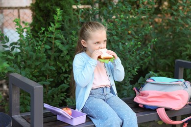 Photo of Cute little girl eating sandwich on bench outdoors. School lunch