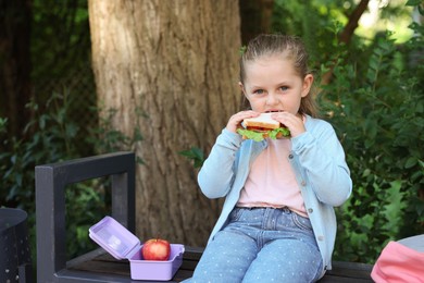 Photo of Cute little girl eating sandwich on bench outdoors. School lunch